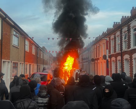 A crowd watch a fire in the middle of a residential street
