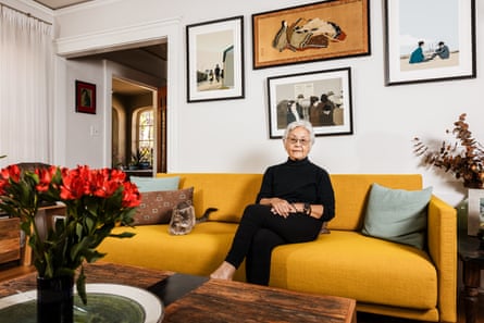 Woman in all black seated on ochre couch with art on wall behind her and bright orange flowers in a vase.