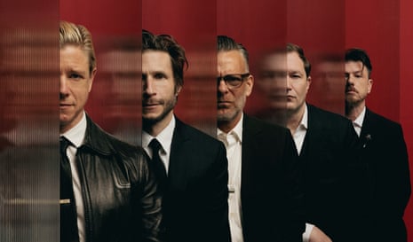 Five men wearing black jackets and red ties, standing in front of a red background