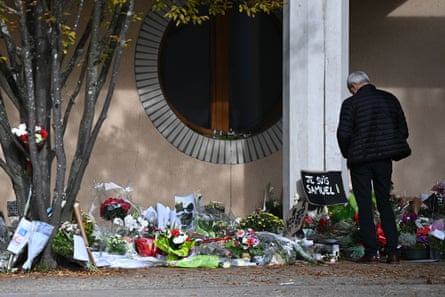 A man looks at flowers laid outside a school building, with a sign saying ‘Je suis Samuel’ visible