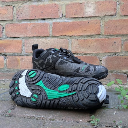 A pair of Saguaro Free shoes in black with bright green details on the tread. One show is upright and the other shoe in the foreground is displaying the sole. There is a pavement and a brick wall background.