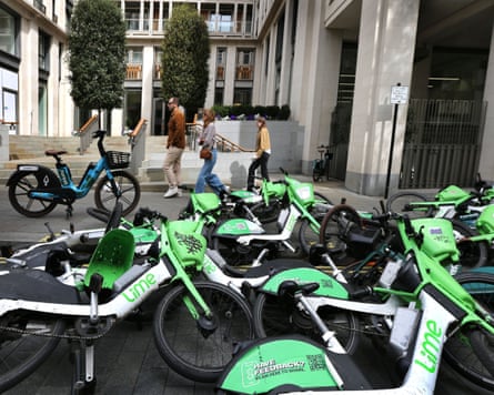 Two women and a man pass in single file in front of a Forest ebike on the pavement and about 10 Lime ebikes lying on top of each other in the street