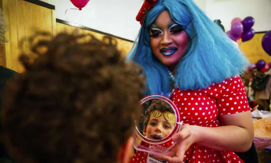 A woman in a red polka dot dress with blue hair and vivid blue makeup holds up a mirror to a child who also has makeup on.