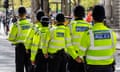 Six Met officers in hi-vis yellow uniform jackets standing in a line on the street, with their backs to the camera