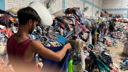 A man holds an item of clothing in a large room filled with piles of clothing