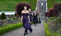 Models walk through along a path between small hedges and purple flowers. Grey stone steps lead down a small grassy slope behind