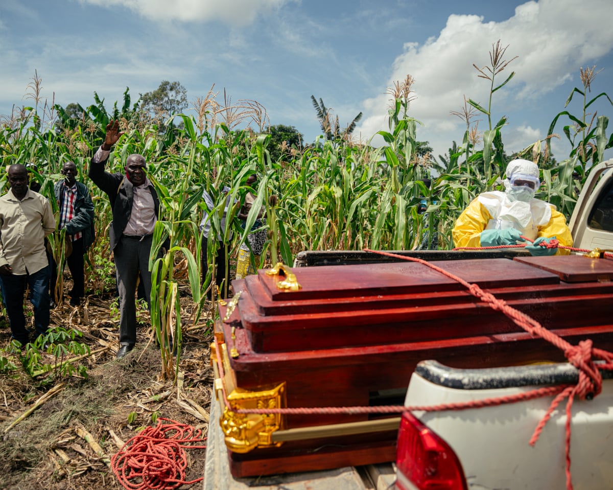 A pastor standing in a cornfield raises his hand as a health worker in a hazmat suit unties a coffin loaded on the back of a pickup truck