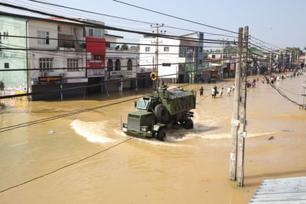 A military vehicle makes its way along a flooded road as people wade through the flood waters behind it