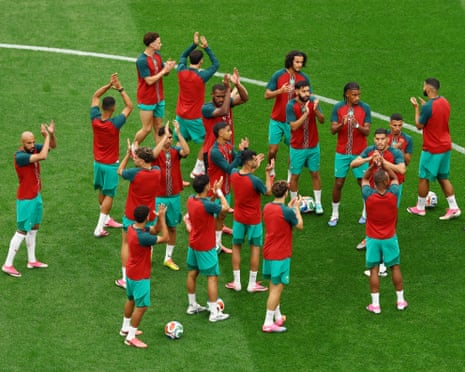 Morocco players in red and teal training kits stand in a circle applauding on a pitch during warmup