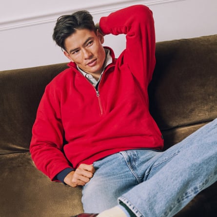 A model wears a bright red J. Crew ‘83 fleece quarter-zip sweatshirt. This is worn with stonewash blue denimjeans and a white under shirt. The model sits on a dark taupe velvet sofa.