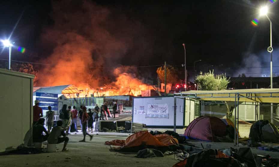 Migrants and riot police stand outside the Moria refugee camp as a fire burns part of the facility.