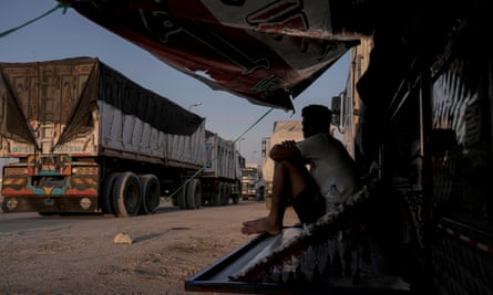 Man sat holding knees in shade with lorry in background.