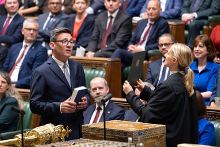 Burnham stands in suit holding book in swearing-in ceremony while MPs look on