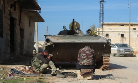 Two rebel fighters sit together behind their tank in a small town with stone houses; one has his back to the camera and sits on a cushion on a stone block, while the other sits on blankets on the ground. They appear to have made a small campfire and may be eating or smoking as one man has his hand to his mouth; a tin cup is on the ground in front of him.