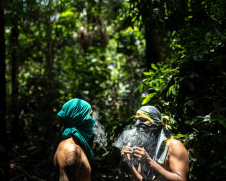 Two bare-chested men with their T-shirts hiding their faces smoke in a jungle clearing