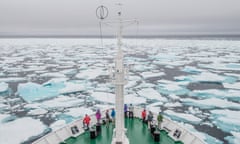 Looking down on the bow of a boat, lined with a few passengers looking out to sea, sailing through ice floes.