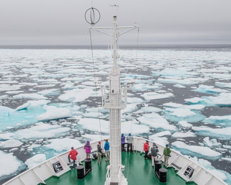 Looking down on the bow of a boat, lined with a few passengers looking out to sea, sailing through ice floes.