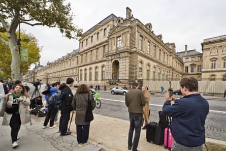 A cluster of people, some with cameras and taking pictures with their phones, stand on the pavement across the road from the stone buildings of the museum.