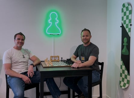 Two men sit at a chessboard, with a neon pawn on the wall, and a chess-themed snowboard