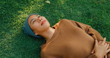 A young Asian woman lies down on grass, taking a moment to relax and breathe in the peaceful atmosphere of sunny park.