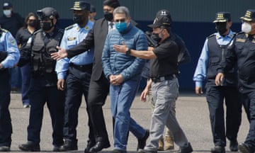 man in handcuffs wearing a blue surgical mask escorted by police officers
