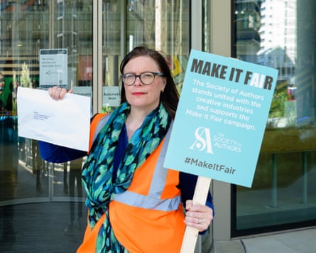 Anna Ganley holds a sign saying Make It Fair. She is wearing an orange hi-vis vest and a green scarf.