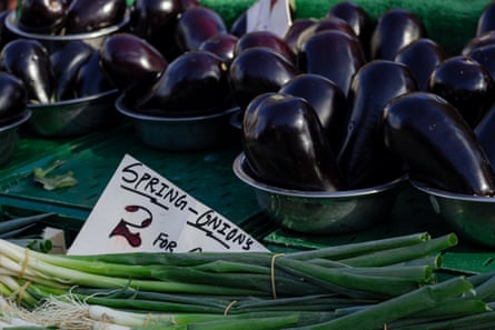 Shiny dark aubergines in metal bowls sit beside bundled spring onions at a market stall
