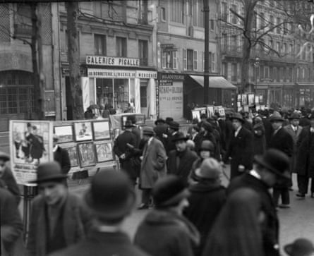 stalls of paintings are set out in front of the buildings as crowds of people in hats and coats walk past.