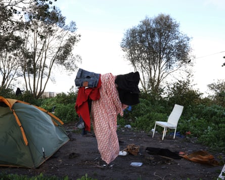 A tent and white chair sit near bushes at a migrant camp, clothes hanging to dry