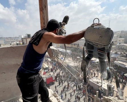 A man standing on the fourth floor of a damaged building holds up a TV camera and rucksack to show a crowd of people in the street below