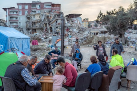 People sitting outside at a table amid a cluster of tents with destroyed buildings in the background