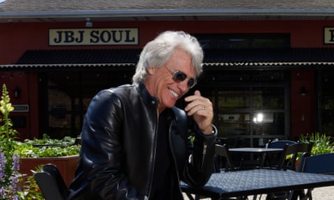 Jon Bon Jovi, wearing leather jacket and sunglasses, smiles as he looks down into his hand.