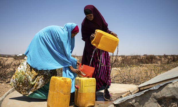 Women in an arid landscape fill plastic containers with water at a well
