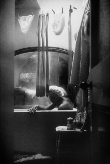 A woman in a shower cap rests in a bathtub in a shared London flat, seen through hanging laundry
