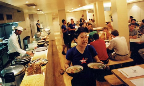 The brightly lit inside of the first ever Wagamama in London’s Bloomsbury, the kitchen with chefs busy to the left and customers seated on benches and stools at tables in the background, a waitress carrying two dishes and smiling broadly in the foreground