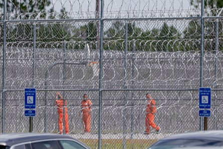Three men in orange jumpsuits stand outside by wire fencing