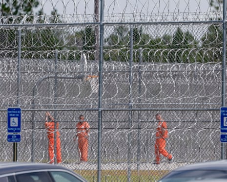 Men in orange jumpsuits behind a high chain-link fence laced through with razor wire.