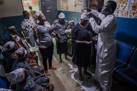 A group of pregnant women sit and stand around at a clinic, while a man attends to one woman’s head
