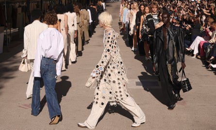 A line of models walk in a circle. Focus of the image is on a person wearing a long dress-like garment that has polka dots made of small mirrors 