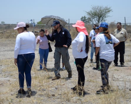 A man gestures as he walks across on dry, rocky soil and talks to seven women as another man looks on