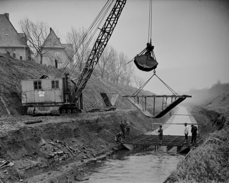A black and white photograph shows a crane lower a concrete shell into a waterway