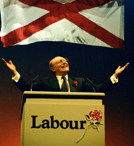 Neil Kinnock holding his arms out while speaking at a yellow podium with Labour and a red rose on it, with a white flag with a red saltire on it behind him