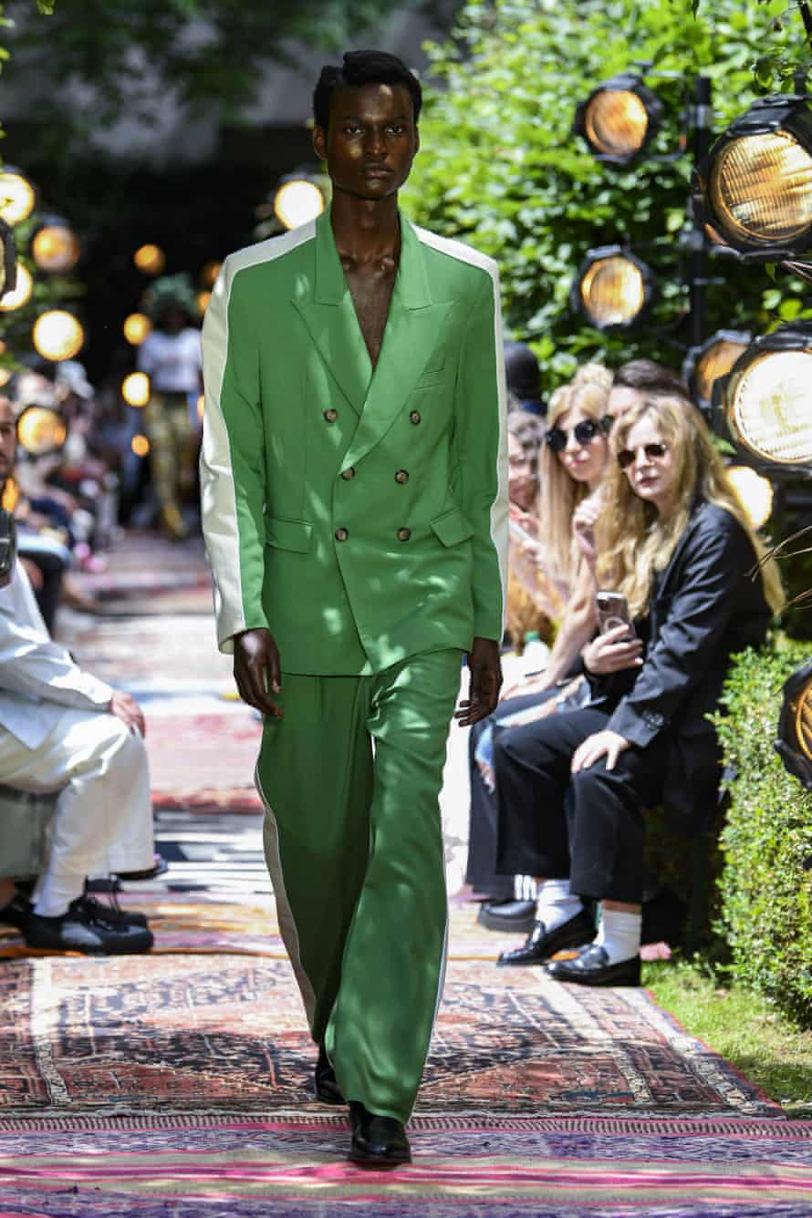 A Black male model is walking down a catwalk wearing a green double-breasted suit with white shoulders and sleeves.
