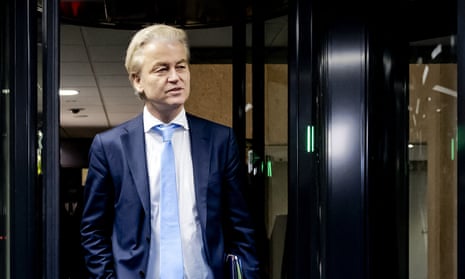 Geert Wilders walks through doorway by a security scanner and camera, and is wearing a dark blue suit with jacket open, white shirt and pale blue tie