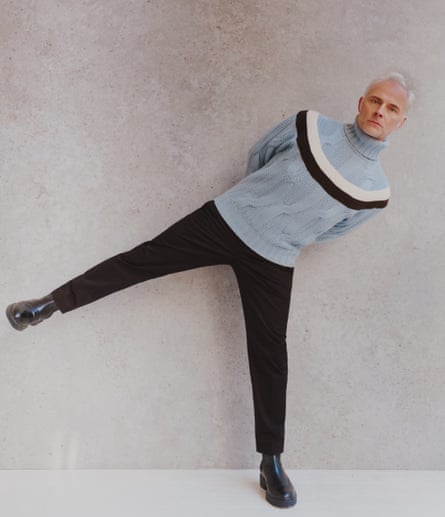 Mark Bonnar, standing on one leg, the other straight and out to the side, wearing pale blue cable knit with a ring of dark blue and white, dark trousers and boots, all hermes.com
