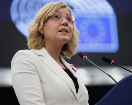 A woman with blond hair wearing a white blazer speaks at a lectern to the backdrop of an EU flag.