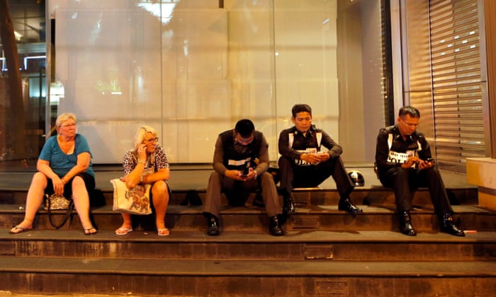Two Western tourists sit next to Thai police officers using their mobile phone at a closed shopping mall near the bomb outside the Erawan Shrine in central of Bangkok
