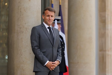 Emmanuel Macron stands outside the Élysée Palace with his hands clasped in front of his body. He wears a grey suit and dark tie.