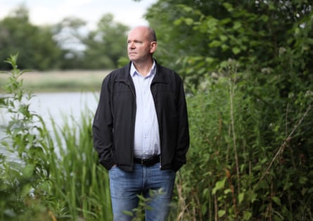 A man in jeans and a black jacket standing among bushes with a lake behind him
