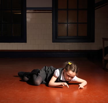 A girl in a short-sleeved white shirt and grey school tunic is lying on the floor and appears to be playing with something small